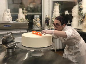 Madison Lee cake designer leveling fondant on the bottom tier of a wedding cake. Dummy cakes in the background with beautiful sugar flowers. Chef Madison Lee wears her iconic red glasses and printed headband over her chef coat.